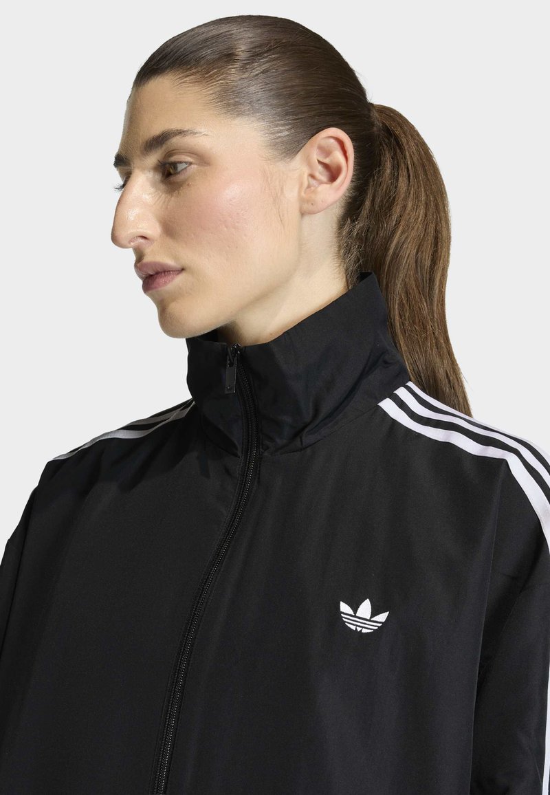 Donna con coda di cavallo marrone che indossa una giacca nera Adidas con strisce bianche e logo, rivolta verso sinistra su uno sfondo chiaro uniforme.
