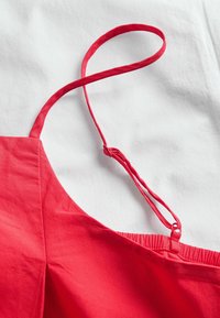 Rød ærmeløs beklædningsgenstand med justerbar tynd strop og elastisk detalje på bagsiden på hvidt stofbaggrund.