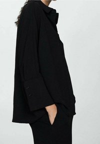 Chemise noire oversize en tissu texturé, dotée d'un col classique, de manches longues avec poignets à boutons et d'un détail de poche latérale.