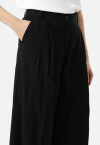 Schwarze weite Hosen aus Stoff mit glatter Textur. Verfügen über einen vorderen Knopfverschluss und Seitentaschen. Maßgeschneidertes Design mit Falten.