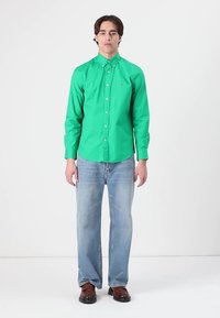Camicia verde con colletto, abbinata a jeans dritti blu chiaro e scarpe marroni, su uno sfondo semplice.
