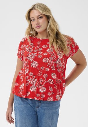 Woman with long blonde hair wearing a red floral short-sleeve blouse and blue jeans, posing with one hand on her hip.