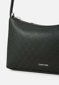Juodas įspaudas rankinukas su geometriniu raštu, turintis užtrauktuką ir petinę juostą, pažymėtas sidabriniu "Calvin Klein" logotipu.