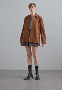 Hnědá suédová oversized bunda s knoflíkovým zapínáním, šedý top, fialová vzorovaná sukně a černé kožené boty až ke kolenům.