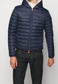 Marineblauwe gewatteerde jas met een doorgestikt ontwerp, capuchon en een rits aan de voorkant. Voorzien van een oranje ritslip en geribde manchetten. Draagt over donkere jeans.