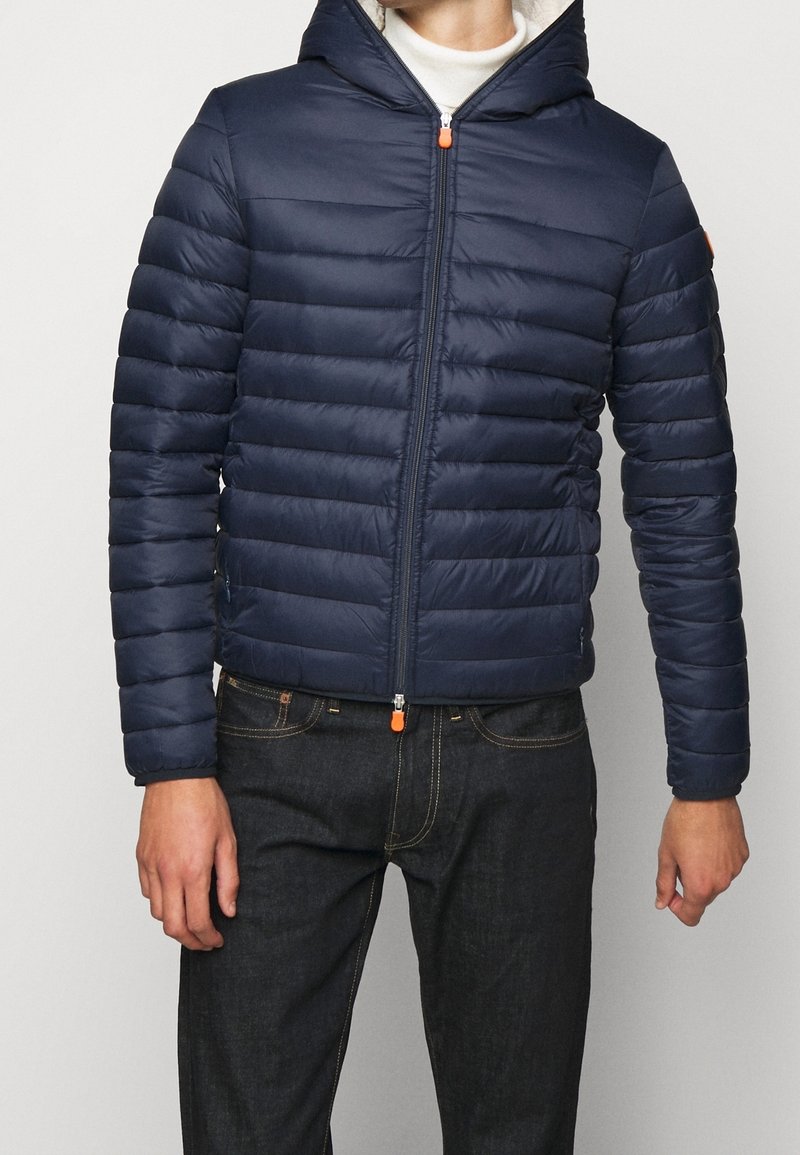 Marineblauwe gewatteerde jas met een doorgestikt ontwerp, capuchon en een rits aan de voorkant. Voorzien van een oranje ritslip en geribde manchetten. Draagt over donkere jeans.