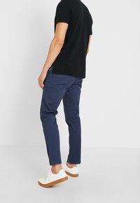 T-shirt nero a maniche corte, pantaloni slim-fit blu navy e sneakers bianche con suole in gomma marrone. Il T-shirt è leggermente più lungo sul retro.