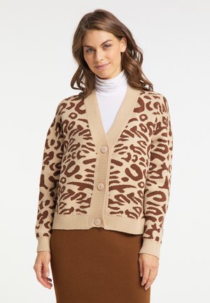 Frau trägt eine beige-braune Leopardenmuster-Strickjacke über einem weißen Rollkragenpullover und einem braunen Rock, steht vor einem einfarbigen Hintergrund.