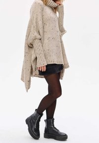 Beige oversized knit sweater with a high collar, textured pattern, and side slits, paired with a denim skirt and black lace-up ankle boots.