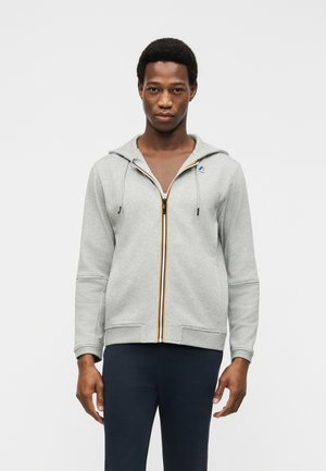 Νέος άνδρας που φοράει ανοιχτό γκρι φούτερ με φερμουάρ και μικρό λογότυπο στο στήθος, συνδυασμένο με σκουρόχρωμο παντελόνι, στέκεται μπροστά σε απλό φόντο.