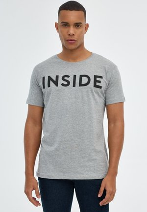 Hombre con cabello corto de pie mirando hacia adelante, vistiendo una camiseta gris con la palabra "INSIDE" en letras negras y en negrita, y pantalones azul oscuro.