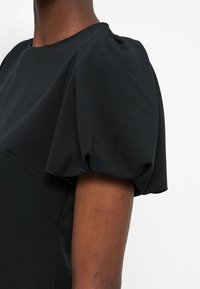 Άτομο που φοράει μαύρο τοπ με κοντά φουσκωτά μανίκια, εμφανίζοντας τον ώμο και το άνω μέρος του βραχίονα σε λευκό φόντο.