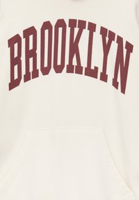 Sweat à capuche blanc avec un grand texte bordeaux « BROOKLYN » en arc sur la poitrine et une poche kangourou visible à l'avant en bas.