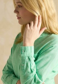 Mintgroene blouse met een ruitpatroon, lange mouwen, knoopmanchetten en geplisseerde details bij de polsen. Gladde stoftextuur.