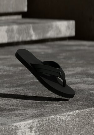 Fekete flip-flop szandál texturált betonlépcsők felett lebeg, árnyékot vet a természetes fényben a talajra.