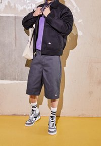 Svart jacka, lila tröja, grå shorts och grå sneakers med vita detaljer. Texturen varierar mellan materialen, vilket ger kontrast i designen.