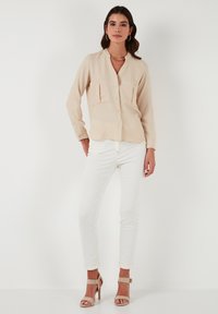 Beige button-up blouse with two front pockets, long sleeves, and a relaxed fit, worn with white fitted pants and strappy beige heels.
