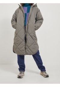 Abrigo largo gray acolchado con capucha, con cierre de cremallera, bolsillos laterales y un patrón geométrico. Combinado con jeans azules y zapatillas.