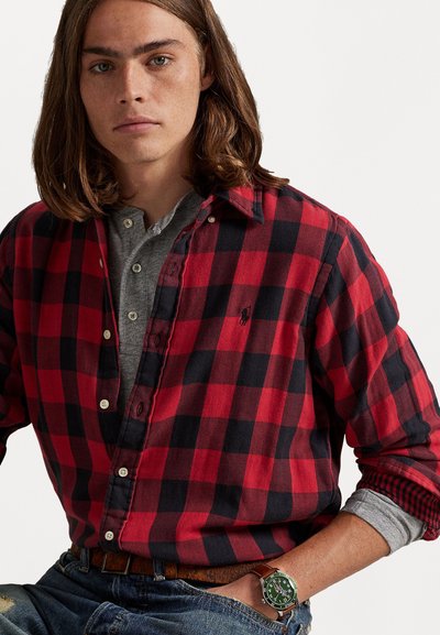 Κόκκινο και μαύρο καρό φανελένιο πουκάμισο με κλειστό γιακά, τοποθετημένο πάνω από γκρι χενλέι. Ορατό ρολόι με �πράσινο καντράν.