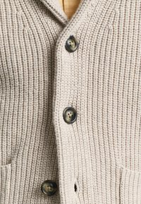 Krämfärgad stickad cardigan med mörka knappar och synliga framsidor, som visar ett strukturerat ribbmönster.