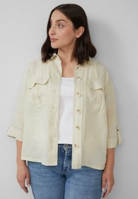 Lichtbeige linnen shirt met knoopsluiting, twee borstzakken en opgerolde mouwen, gecombineerd met een witte tanktop en blauwe spijkerbroek.