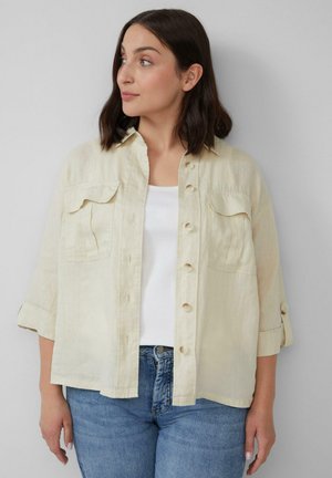 Lichtbeige linnen shirt met knoopsluiting, twee borstzakken en opgerolde mouwen, gecombineerd met een witte tanktop en blauwe spijkerbroek.