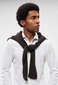 Pruun kootud sall, mis on seotud kaela ümber valge särgi peale. Sallil on tekstuuriga kangas ja lahtine, vaba sõlm.