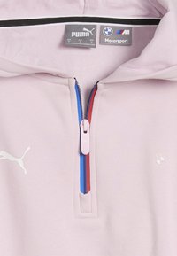 Hele roosa pullover, millel on tõmblukuga kaelus, esitatuna sinise ja punase triibuga tõmbluku ääres. Sellel on valge Puma logo ja BMW Motorsport kaubamärk.