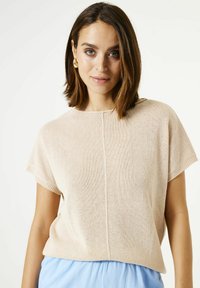 Vrouw met schouderlang bruin haar, dragend gouden oorbellen, een beige kortmouw gebreid shirt en lichtblauwe broek, staand tegen een witte achtergrond.