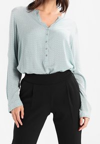 Chemisier bleu clair à motifs avec un devant à boutons et un décolleté en V, associé à un pantalon noir taille haute. Tissu doux avec des manches longues.