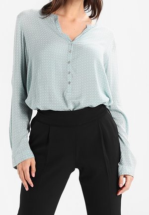 Lichtblauwe blouse met patroon, met een knoopsluiting aan de voorkant en een v-hals, gecombineerd met hoge zwarte broek. Zacht materiaal met lange mouwen.