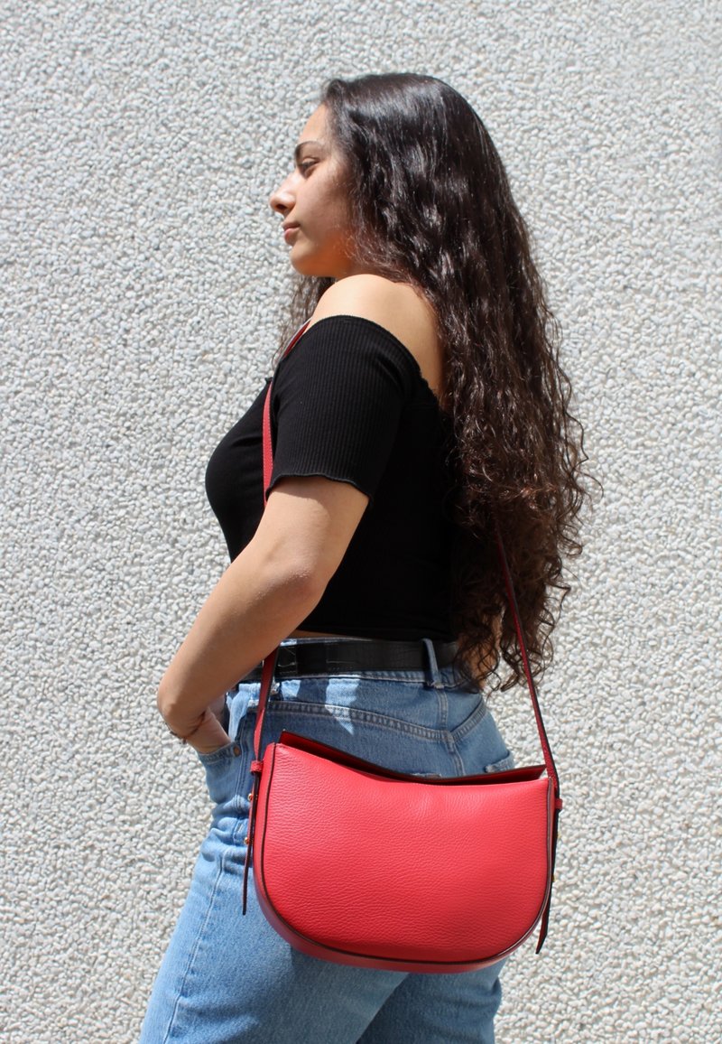 Bolso de hombro de cuero rojo con forma curva, acabado texturizado y una única correa ajustable, llevado en la cadera sobre un fondo de mezclilla.