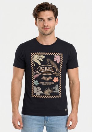 Hombre con camiseta negra de Von Dutch con estampado colorido de hojas y flores tropicales y jeans azules, de pie frente a un fondo blanco.