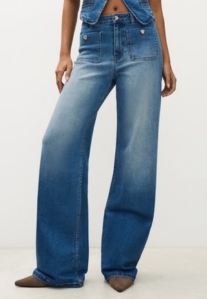 Jean bleu taille haute à jambes larges avec poches plaquées à l'avant et boutons, porté avec des chaussures marron à bout pointu et un top en denim court.