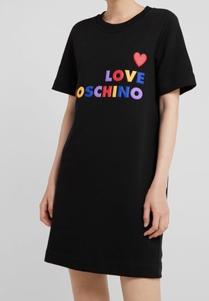 Zwarte jurk met korte mouwen met kleurrijke "LOVE MOSCHINO" letters en een rood hart op de borst, gedragen door een staand persoon.