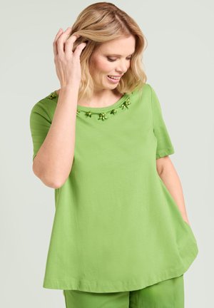 Donna con capelli biondi, indossa una maglietta verde a maniche corte con scollo decorato con perline, sorride e si tocca i capelli con una mano, su uno sfondo neutro.