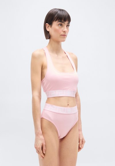 Γυναίκα με ανοιχτό ροζ αθλητικό σουτιέν και σλιπ ψηλής μέσης με διακριτικό γεωμετρικό μοτίβο  στη μέση, στέκεται μπροστά σε απλό ανοιχτό φόντο.