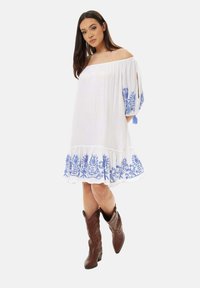 White off-the-shoulder dress with blue floral embroidery at the sleeves and hem; textured fabric; paired with brown cowboy boots.