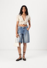 Beige lyhyt cropped-paita, jossa on puffihihat ja pitsireunus, yhdistettynä vaaleansinisiin denim-shortsihin. Malli pitää kädessään pientä valkoista käsilaukkua ja käyttää mustia balettikenkkiä.