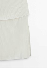 Top in tessuto a coste di colore crema con design a strati e una spaccatura laterale per facilitare i movimenti. Texture morbida con un leggero luccichio.