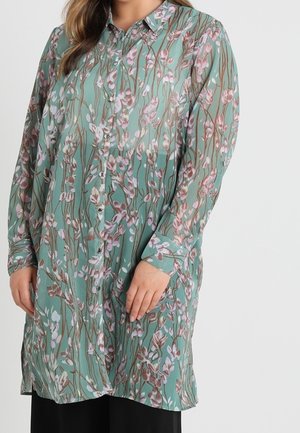 Mujer con túnica azul verdosa de manga larga con botones, con un estampado floral rosa y marrón, combinada con pantalones negros.