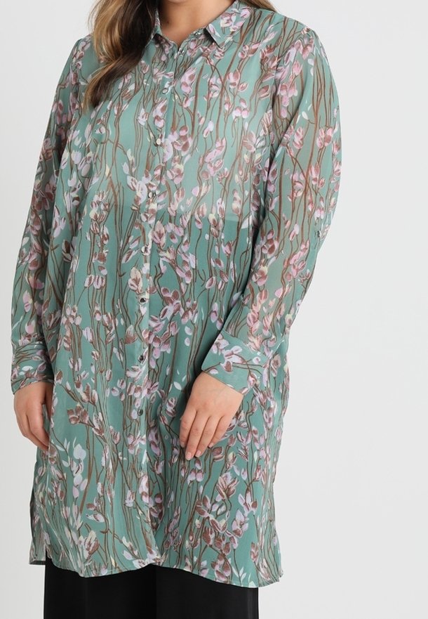 Femme portant une tunique bleu sarcelle à manches longues, boutonnée, avec un motif floral rose et marron, associée à un pantalon noir.