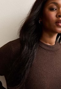 Suéter de punto marrón con cuello redondo, que presenta un acabado texturizado. El modelo tiene el cabello largo y ondulado de color oscuro y lleva un pequeña aro en una de las orejas.