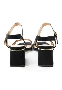 Zwarte suède heeled sandals hebben een blokhak met een gouden accent, een verstelbare enkelriem en een open teenontwerp. Zachte binnenvoering zichtbaar.
