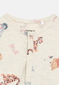 Kreemivärvi beebimüts, millel on nööbid ja millel on väiksed printsid Notsu, Tunne ja Karu Tuhkatriinu tegelastest.