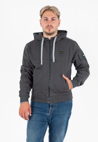 Felpa grigia con zip, cappuccio regolabile e tasche frontali, con una toppa sul braccio sinistro. Realizzata in un morbido tessuto testurizzato. Vestibilità casual.