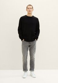 Suéter negro de punto, jeans grises y zapatillas blancas. La persona tiene una expresión neutral, de pie contra un fondo claro.