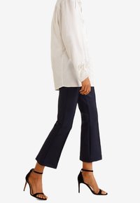 Zwarte cropped broeken met een op maat gemaakte pasvorm, gecombineerd met een witte blouse met knopen en zwarte, strappy hoge hakken. Glad stoffen, enkelbandjes zichtbaar.