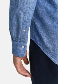 Lichtblauw linnen shirt met een knoopmanchet, met een subtiele textuur en een gebogen zoom, getoond tegen een donkere broek achtergrond.