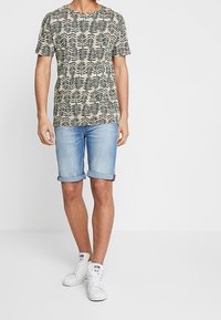 Beige t-shirt med kort ärm och svart bladmönster, tillsammans med ljusblå denimshorts och vita gymnastikskor. Lätt sommaroutfit.